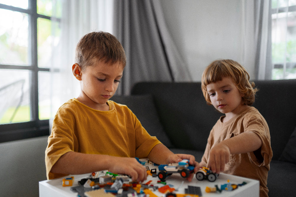 Two children playing with LEGO on a table in a room with large windows.